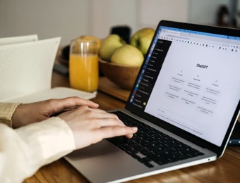 Hände tippen auf einem Laptop auf einem Holztisch; daneben Obstkorb, Glas Orangensaft und ein Notizblock.