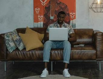 Mann sitzt auf braunem Ledersofa, Laptop im Schoß; Wand mit orangefarbener Kunst, mehrere Kissen.