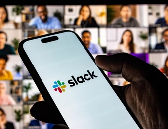 Eine Hand hält ein Smartphone mit dem Slack-Logo; im Hintergrund ein Videokonferenz-Gitter mit vielen Teilnehmern.