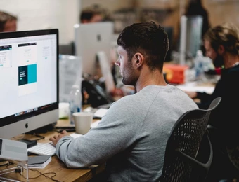 Person sitzt seitlich vor einem großen Computerbildschirm an einem Schreibtisch; Hintergrund mit weiteren Arbeitsplätzen.