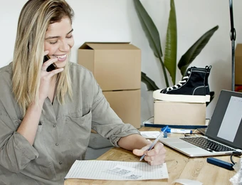 Person sitzt am Schreibtisch, telefoniert, schreibt; Laptop, Schuh auf Box, Kartons, Pflanze im Hintergrund.