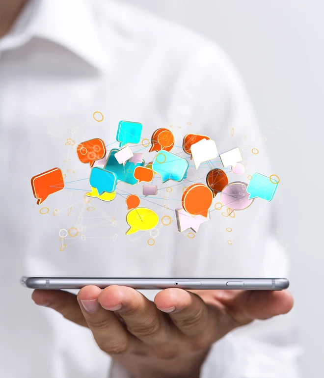 Hände halten ein silbernes Tablet; bunte Sprechblasen-Symbole in Orange, Blau und Gelb schweben darüber.