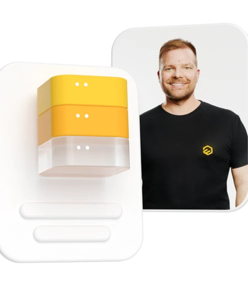 Gelb-weißes Gerät im Vordergrund vor weißem Hintergrund; rechts steht ein Mann in schwarzem T‑Shirt.