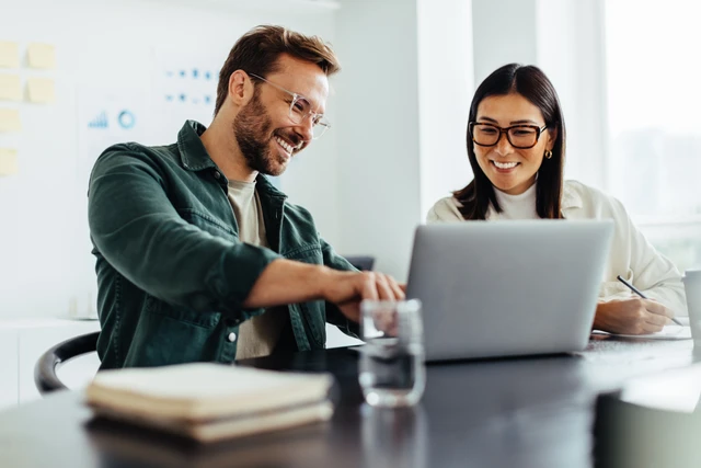Zwei lächelnde Personen sitzen an einem Tisch mit Laptop; Wasser, Notizbuch, helles Büro im Hintergrund.
