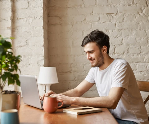 Junger Mann arbeitet an einem Laptop am Holztisch; Becher, Notizblock, Pflanze und Lampe, weiße Backsteinwand im Hintergrund.