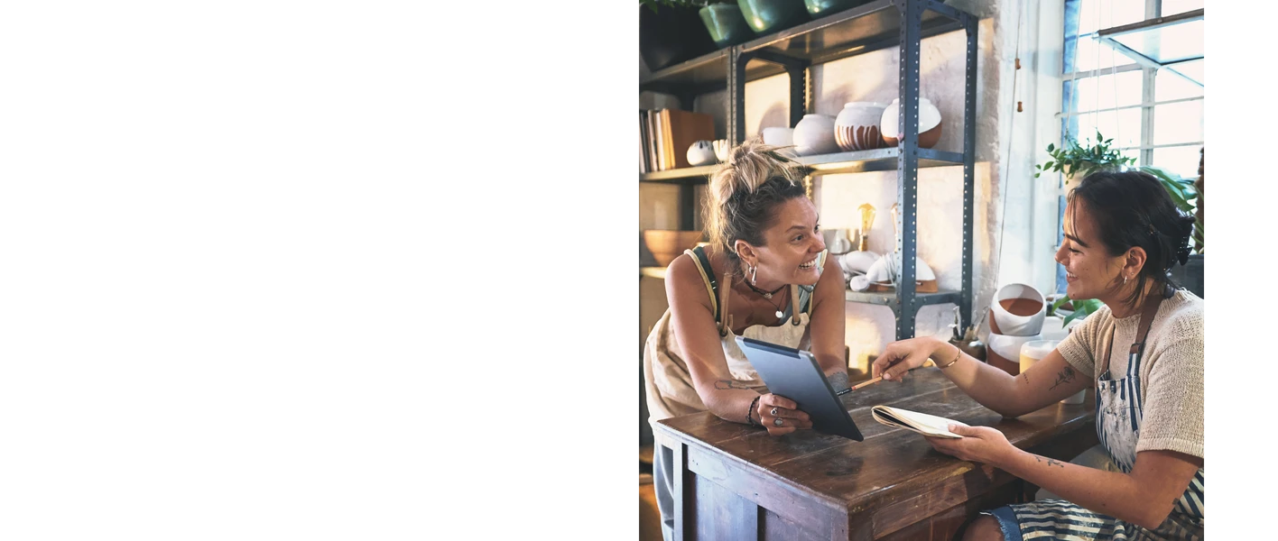 Zwei Frauen lehnen an einem Holztresen, eine hält ein Tablet, die andere reicht einen Teller; Regal mit Geschirr im Hintergrund.