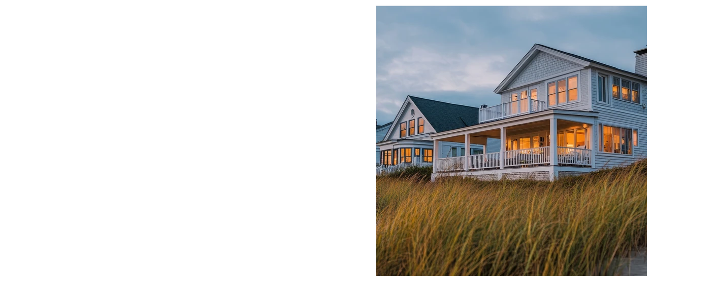 Zweistöckiges Haus mit Veranda und vielen Fenstern, Dünengras im Vordergrund, blauer Himmel.