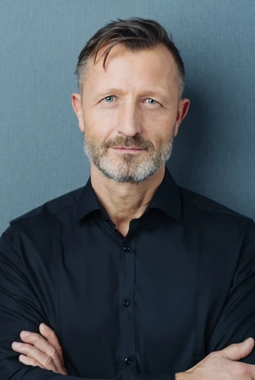 Mann mittleren Alters mit kurzem dunklem Haar, grauem Bart, schwarzem Hemd, Arme verschränkt, vor blauem Hintergrund.