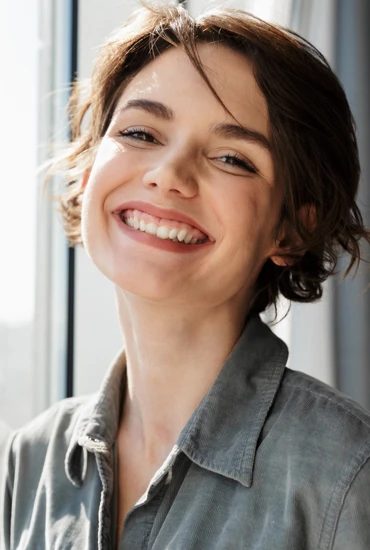 Frau mit kurzen braunen Haaren, breites Lächeln, graues Hemd, steht vor einem Fenster.