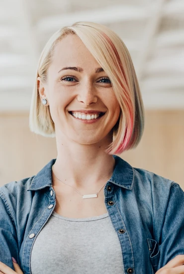 Lächelnde Frau mit kurzem blondem Bob, pinke Strähne, Jeanshemd über grauem Top, Ohrringe und rechteckige Kette; heller Innenraum