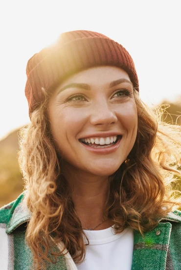 Lächelnde Frau mit lockigen Haaren, roter Strickmütze, grün-karierte Jacke und weißem Oberteil; Sonnenlicht von links.
