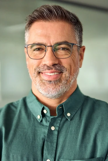 Mann mittleren Alters mit grauem Bart und Brille, grünem Hemd, lächelnd, unscharfer Hintergrund.