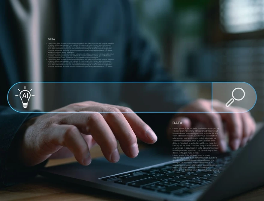 Nahaufnahme: Person tippt auf Laptop; holografische Suchleiste mit AI-Icon links, Lupe rechts.