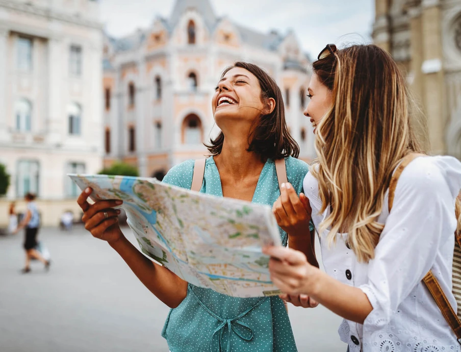 Zwei Frauen halten eine Karte; eine lacht, im Hintergrund Gebäude in einer Stadt.