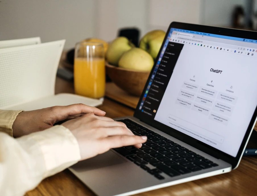 Hände tippen auf einem Laptop auf einem Holztisch; daneben Obstkorb, Glas Orangensaft und ein Notizblock.