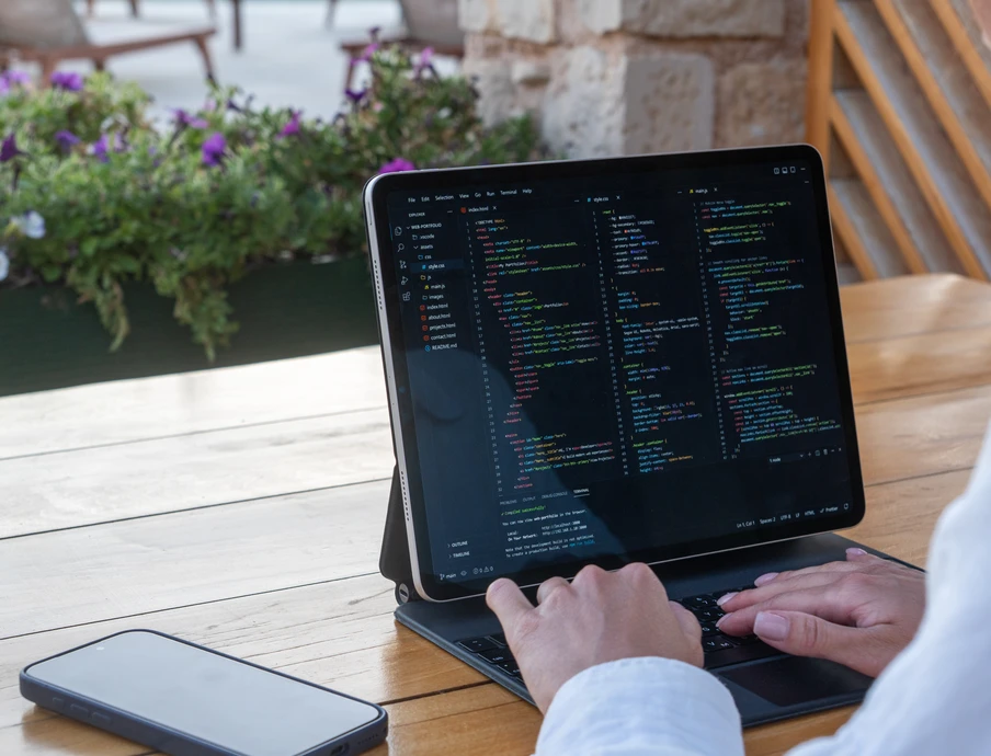 Person tippt auf Laptop mit Codeanzeige; Smartphone daneben, Holztisch im Freien, Blumen im Hintergrund.