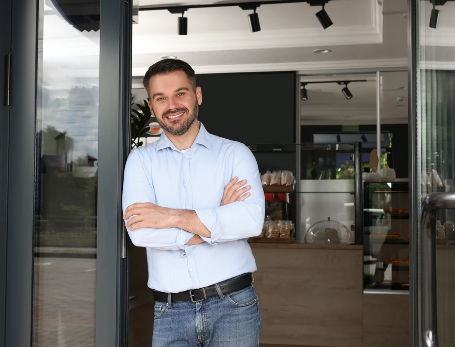 Mann in hellblauem Hemd und Jeans steht mit verschränkten Armen in offener Glastür eines Innenraums und lächelt.
