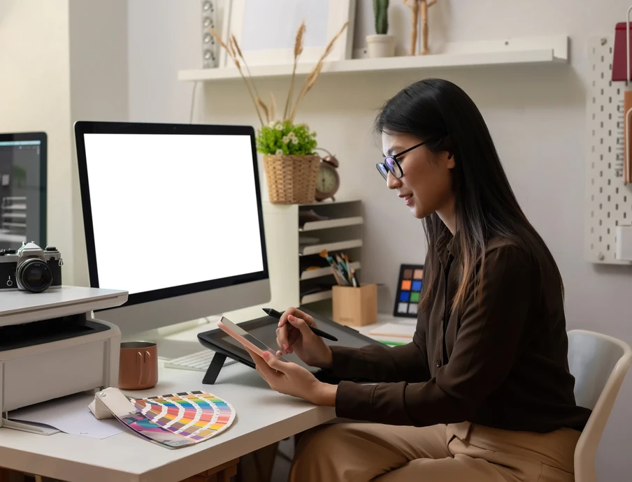 Frau sitzt am Schreibtisch, hält Tablet und Stift, vor Monitor mit weißem Bildschirm; Farbfächer und Kamera auf dem Tisch.