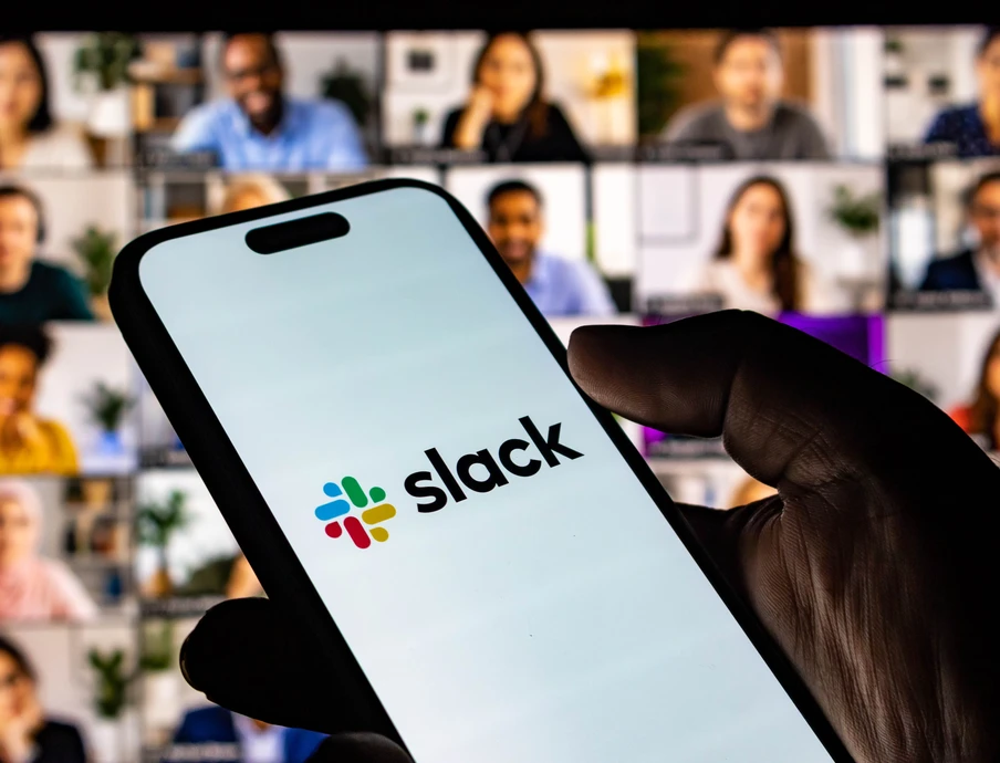 Eine Hand hält ein Smartphone mit dem Slack-Logo; im Hintergrund ein Videokonferenz-Gitter mit vielen Teilnehmern.