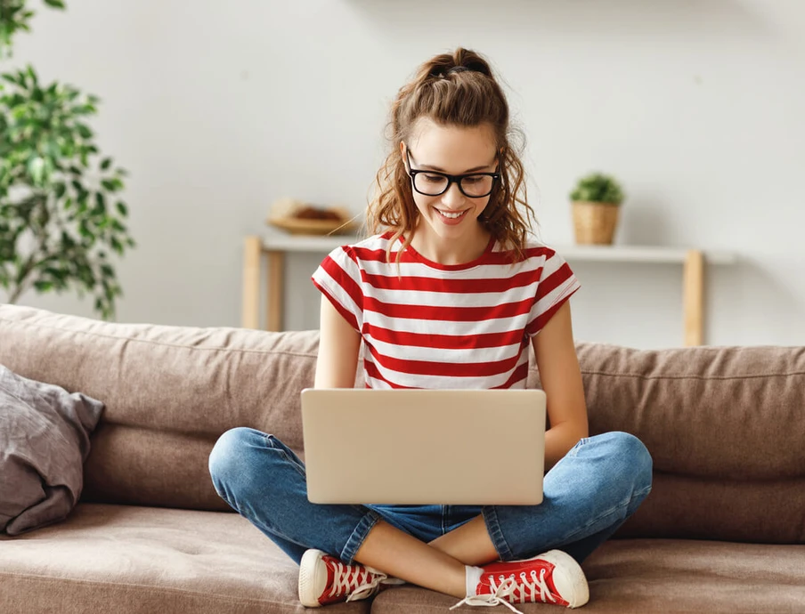 Frau sitzt auf braunem Sofa, Laptop im Schoß, rote/weiße Streifenbluse, Jeans, rote Sneaker, Brille; Pflanze im Hintergrund.