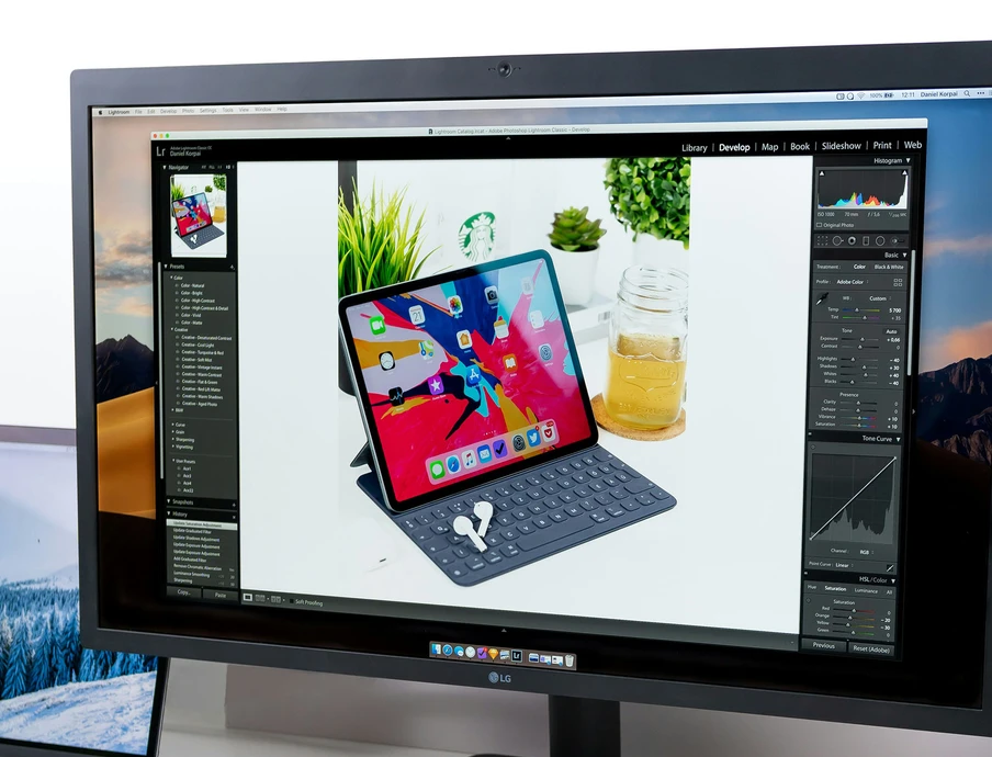 Monitor zeigt Fotobearbeitung, darauf ein iPad mit Tastaturhülle und Stift auf weißem Tisch, Pflanze im Hintergrund.
