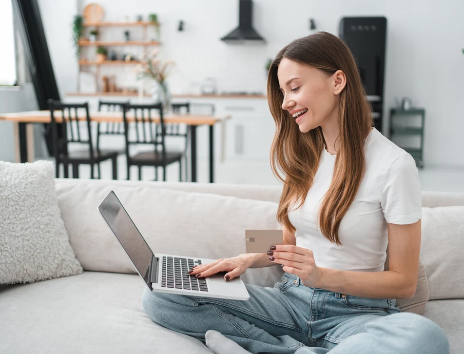 Frau sitzt lächelnd auf dem Sofa, Laptop auf dem Schoß, hält Kreditkarte; im Hintergrund Küche.