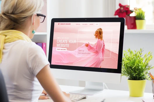 Person sitzt vor dem Computerbildschirm, auf dem eine pinkfarbene Website zu sehen ist; Tastatur, Maus und Pflanze.