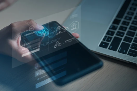 Person hält Smartphone; auf dem Display erscheinen hologrammartige Icons, Laptop im Hintergrund.