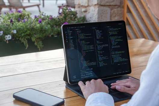 Person tippt auf Laptop mit Codeanzeige; Smartphone daneben, Holztisch im Freien, Blumen im Hintergrund.