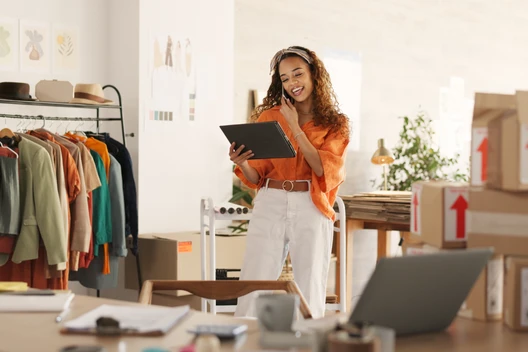 Eine Frau in Orange und Weiß hält ein Tablet; im Hintergrund Kleiderständer, Kartons und Laptop.