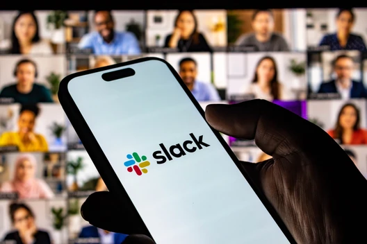 Eine Hand hält ein Smartphone mit dem Slack-Logo; im Hintergrund ein Videokonferenz-Gitter mit vielen Teilnehmern.