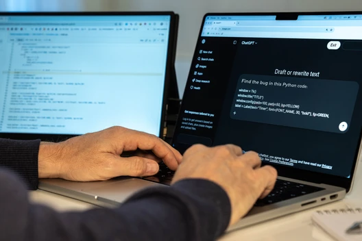 Person tippt auf einem Laptop; zwei Monitore im Hintergrund zeigen Code und ein Chatfenster.