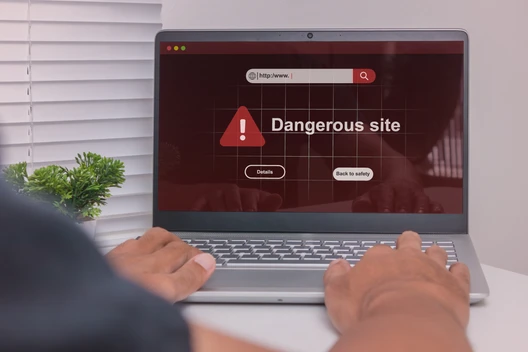 Laptop auf weißem Tisch; Bildschirm zeigt Warnung Dangerous site mit rotem Dreieck, Hände auf der Tastatur.