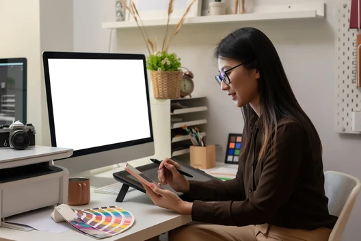 Frau sitzt am Schreibtisch, hält Tablet und Stift, vor Monitor mit weißem Bildschirm; Farbfächer und Kamera auf dem Tisch.