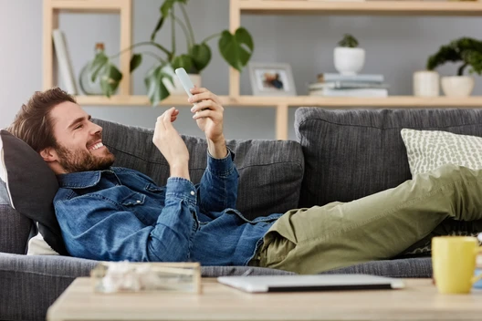 Mann liegt entspannt auf grauem Sofa, schaut auf Smartphone, trägt Jeansjacke und grüne Hose; Wohnzimmer mit Pflanzen.